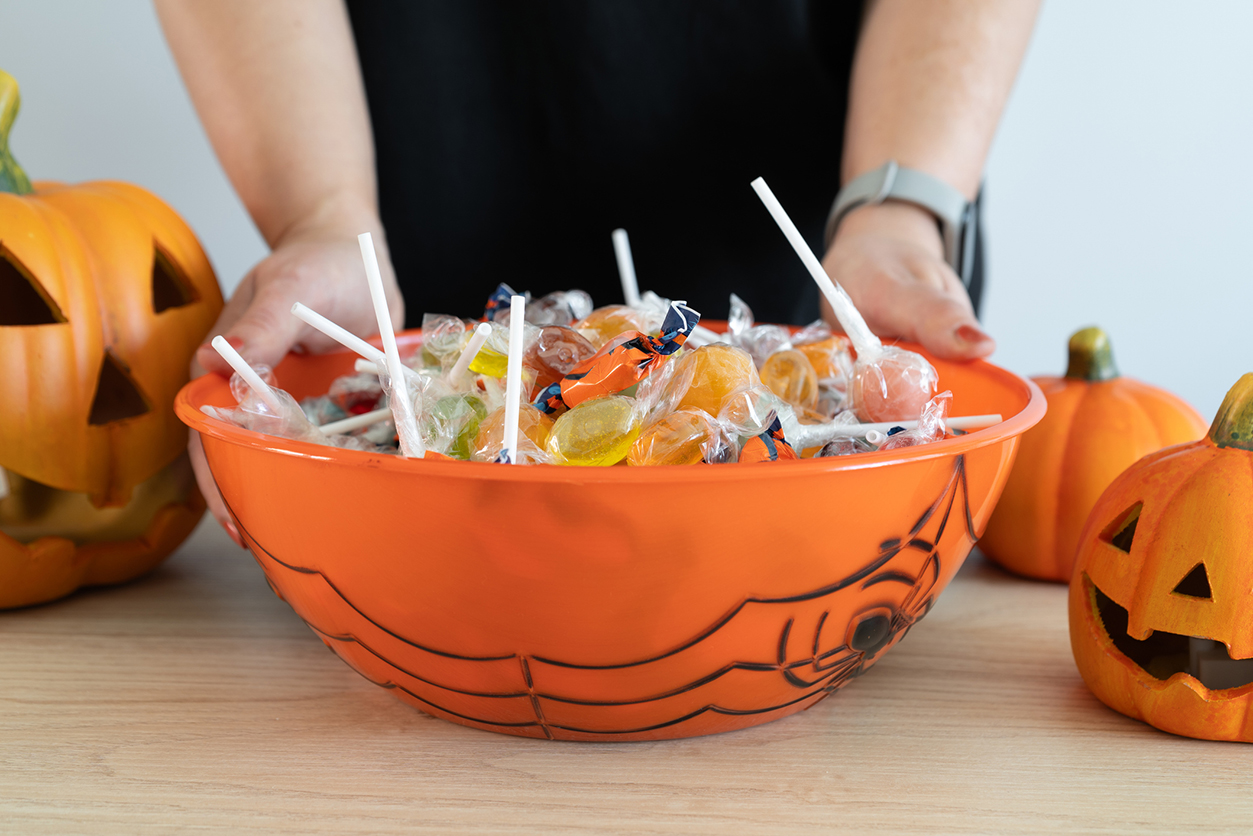 Post-Halloween Dental Check-Up - Woman holding a Halloween candy bowl filled with sweets for trick-or-treating. Female hands and Halloween-themed decorative treat bowl container. Jack-o'-lantern pumpkins placed around the table.