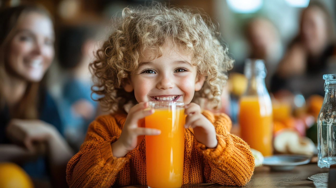 Smiling child with curly hair holding a glass of orange juice at the breakfast table