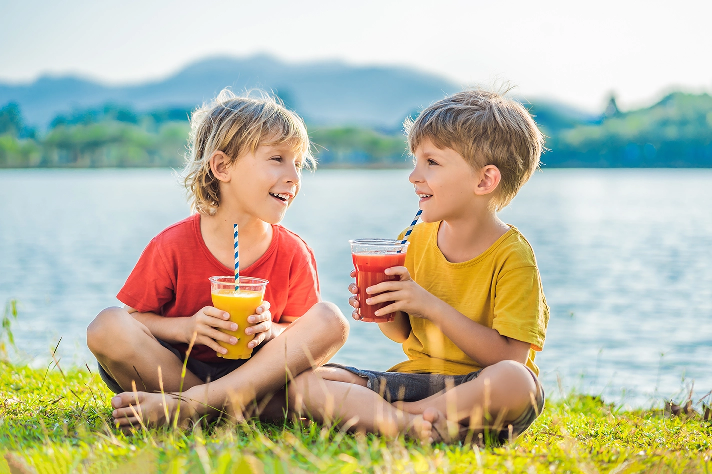 wo young boys sitting on grass drinking orange and red juice with straws